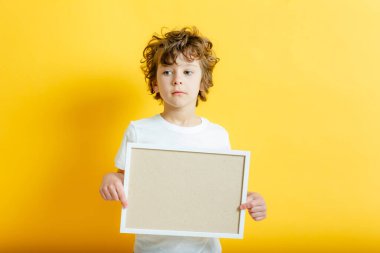 Beyaz tişörtlü, parlak sarı arkaplandaki yazı için boş çerçevenin arkasına saklanan kıvırcık, tatlı çocuk. Bej pano ya da afiş - boşluğu metin doldurmak için kopyala. İleti tahtası