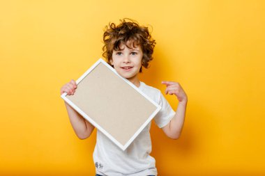 Beyaz tişörtlü kıvırcık çocuk mesaj atmak için boş bir çerçeve tutuyor, gülümsüyor ve parlak sarı arka planda parmağını gösteriyor. Beyaz pano ya da afiş - boşluğu doldurmak için konsepti kopyala. İleti tahtası