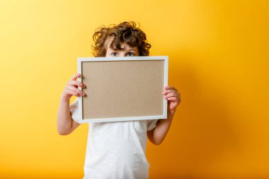 Beyaz tişörtlü, parlak sarı arkaplandaki yazı için boş çerçevenin arkasına saklanan kıvırcık, tatlı çocuk. Bej pano ya da afiş - boşluğu metin doldurmak için kopyala. İleti tahtası