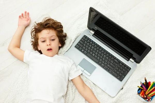 El chico rizado está ocupado con la computadora. colegial mira hacia arriba, ver en la cámara. Concepto de dificultades de la educación en el hogar, el estudio a distancia —  Fotos de Stock