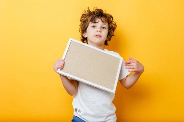 Beyaz tişörtlü kıvırcık çocuk mesaj için boş bir çerçeve tutuyor ve parlak sarı bir arka plan üzerinde parmağını gösteriyor. Beyaz pano ya da afiş - metni doldurmak için boşluk konseptini kopyala. İleti tahtası.