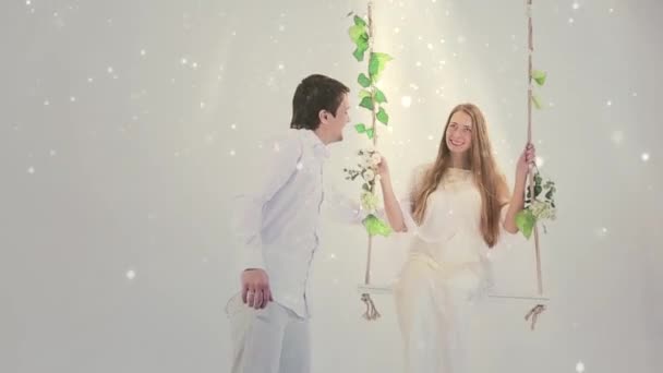 那家伙在一个白色背景的绿色树叶的秋千上摇晃着女孩 他们穿着白色长袍 像亚当和夏娃 太阳照耀着他们 — 图库视频影像