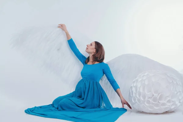 Een zwanger meisje in een blauwe jurk met engelenvleugels op een witte achtergrond. Een engel-vrouw heeft grote witte ballen. Het concept van Kerstmis, mystieke wezens, Halloween — Stockfoto