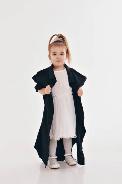 a little girl tries on dad\'s shirt. Child in big black clothes on white background. Concept of children\'s games, imitation of adult life, fashion