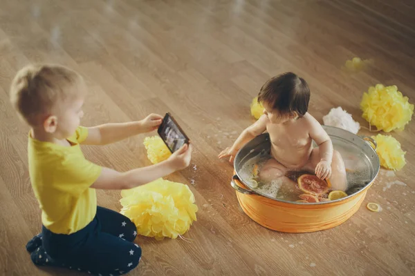 Un niño toma fotos en el teléfono de una niña que se está bañando en un baño con limón y pomelo. Un niño se lava en un lavabo entre flores amarillas. concepto de infancia, cuidado de la salud, FIV, higiene , — Foto de Stock