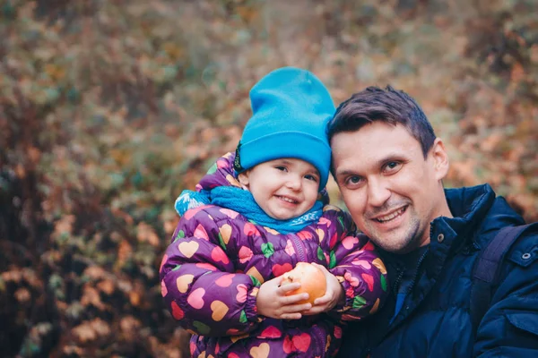 Malé dítě s červeným jablkem s otcem v teplém obleku se prochází lesem. Podzimní park. Koncept dětské módy, doplňky, venkovní procházky — Stock fotografie