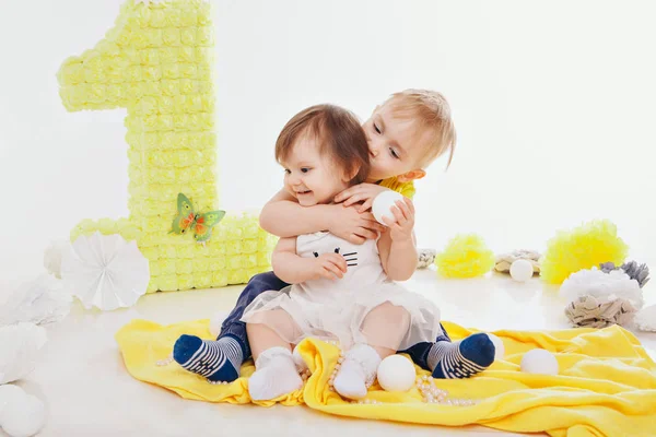 Celebración del cumpleaños: niña y niño sentados en el suelo entre la decoración: números 1, flores artificiales y bolas blancas —  Fotos de Stock