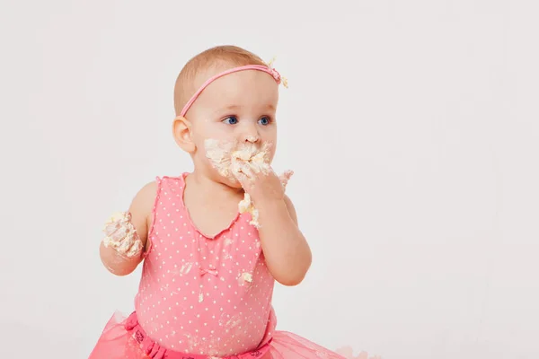 Menina comendo bolo com as mãos no fundo branco. A criança está coberta de comida. Estragou a doçura. O conceito de aniversário, feriados, culinária — Fotografia de Stock
