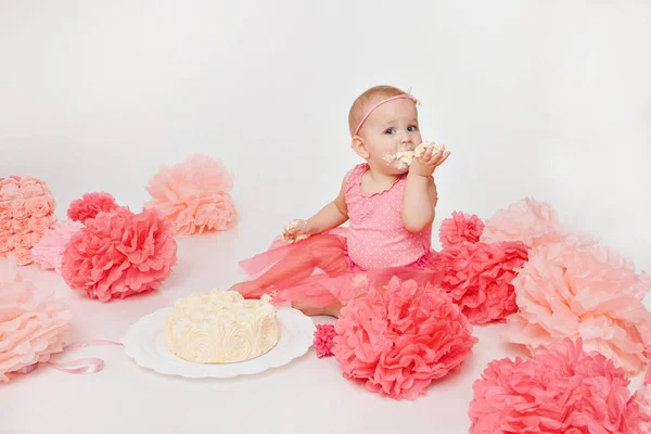 Celebración de cumpleaños: niña comiendo pastel con las manos sobre fondo blanco. El niño está cubierto de comida. Arruinó la dulzura. en el suelo entre la decoración: números 1, flores artificiales a —  Fotos de Stock