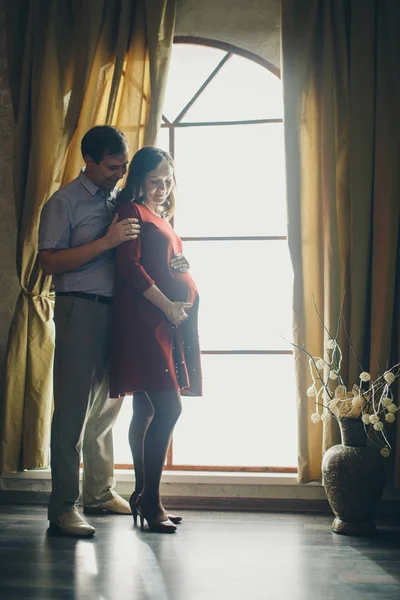 Een liefdevol koppel omarmt aan het raam. Een man en een vrouw kijken elkaar in de ogen. De man knuffelt zijn zwangere vrouw en aait haar maag. Valentijnsdag. 14 februari — Stockfoto