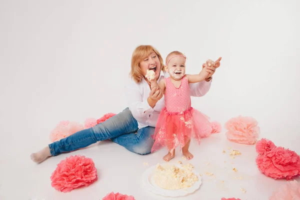 Celebração de aniversário: menina comendo bolo com as mãos no fundo branco. criança está coberta de comida. no chão entre a decoração: números 1, flores artificiais e bolas brancas. Espaço de cópia — Fotografia de Stock