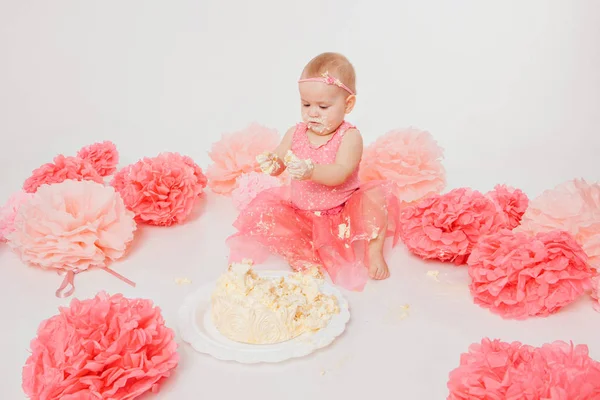 Celebração de aniversário: menina comendo bolo com as mãos no fundo branco. criança está coberta de comida. Doçura arruinada. no chão entre a decoração: números 1, flores artificiais e bolas brancas . — Fotografia de Stock
