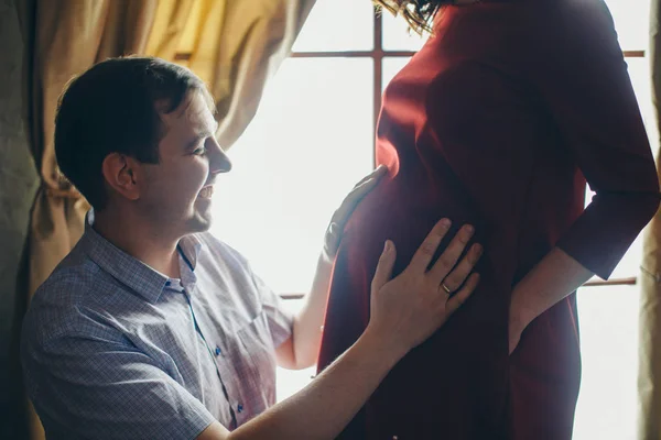 A loving couple embraces at the window. A man and a woman look into each other's eyes. The husband hugs his pregnant wife and strokes her stomach. Valentine's day. February 14 — ストック写真