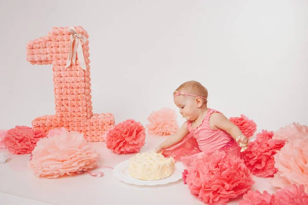 Celebração de aniversário: menina comendo bolo com as mãos no fundo branco. criança está coberta de comida. Doçura arruinada. no chão entre a decoração: números 1, flores artificiais e bolas brancas . — Fotografia de Stock