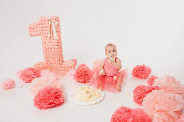 Birthday celebration: little girl eating cake with her hands on white background. child is covered in food. Ruined sweetness. on floor among decoration: numbers 1, artificial flowers and white balls. — Stock Photo, Image