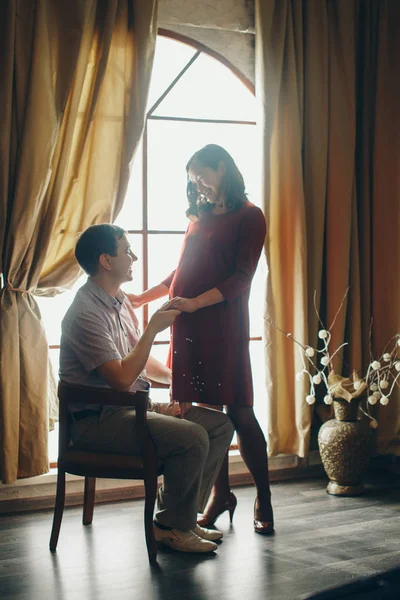 A loving couple embraces at the window. A man and a woman look into each other's eyes. The husband hugs his pregnant wife and strokes her stomach. Valentine's day. February 14 — ストック写真