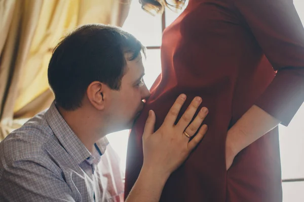 Een liefdevol koppel omarmt aan het raam. Een man en een vrouw kijken elkaar in de ogen. De man knuffelt zijn zwangere vrouw en aait haar maag. Valentijnsdag. 14 februari — Stockfoto