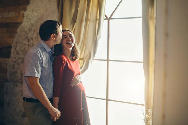 Ett kärleksfullt par famnar vid fönstret. En man och en kvinna ser varandra i ögonen. Mannen kramar sin gravida fru och smeker hennes mage. Alla hjärtans dag. 14 februari — Stockfoto