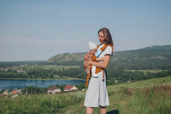 woman with baby in a loose dress with loose hair walks in a meadow. Village houses, forest and river as background. The concept of summer, warmth, freedom, village life, sunburn, childhood