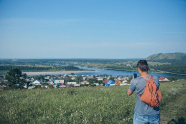 Sırt çantalı bir adam ufku fotoğraflıyor. Bir turist tüm dünyayı gezer. Arka planda köy evleri, orman ve nehir var. Yaz konsepti, sıcaklık, özgürlük, köy hayatı, güneş yanığı
