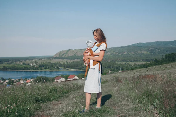 woman with baby in a loose dress with loose hair walks in a meadow. Village houses, forest and river as background. The concept of summer, warmth, freedom, village life, sunburn, childhood