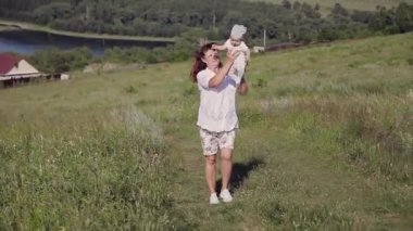Gevşek elbiseli, saçları dökülmüş bir kadın çayırda yürüyor. Köy evleri, orman ve nehir arka planda. Yaz konsepti, sıcaklık, özgürlük, köy hayatı, güneş yanığı, çocukluk