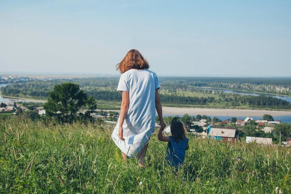 a girl in a loose dress with loose hair walks in a meadow. Village houses, forest and river as background. The concept of summer, warmth, freedom, village life, sunburn