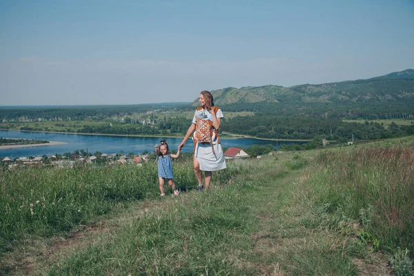 a girl in a loose dress with loose hair walks in a meadow. Village houses, forest and river as background. The concept of summer, warmth, freedom, village life, sunburn