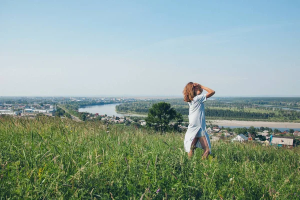 a girl in a loose dress with loose hair walks in a meadow. Village houses, forest and river as background. The concept of summer, warmth, freedom, village life, sunburn