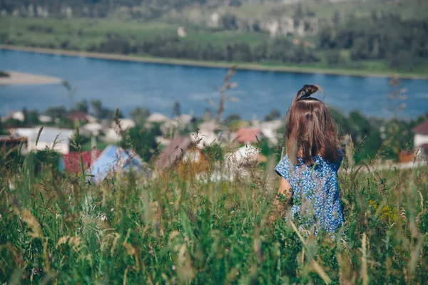 a little girl in a loose dress with loose hair walks in a meadow. Village houses, forest and river as background. The concept of summer, warmth, freedom, village life, sunburn, chldhood