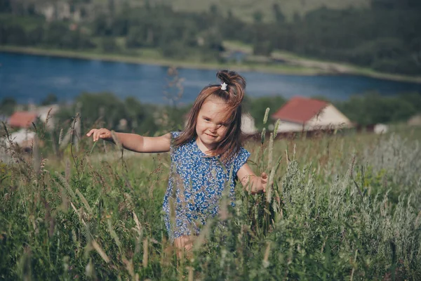 a little girl in a loose dress with loose hair walks in a meadow. Village houses, forest and river as background. The concept of summer, warmth, freedom, village life, sunburn, chldhood