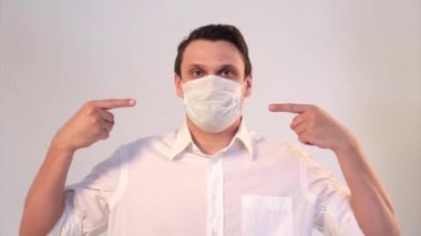 Beyaz arka planda tıbbi maske takan bir adamın portresi. Tıbbi ekipmanların tanıtımı. Adam işaret ediyor. Karantina, kendini izole etme, koronavirüsten koruma, virüs, hastalık korkusu, tedavi, ilaç