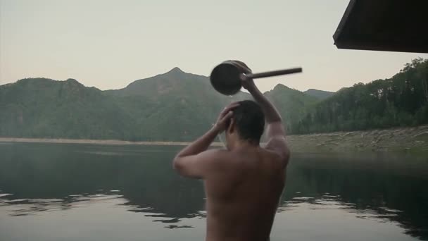 ぬれた男は湖のそばに立つ バケツで水をやってる 冷たい水は体内を流れる 肉体労働の後の幸せな人 体力運動 日焼け 入浴後の体からの蒸気 疲労感 健康的なライフスタイルの概念 — ストック動画