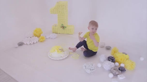 生日庆祝活动 男孩双手拿着白色背景的蛋糕吃饭 孩子身上都是食物 被毁的甜蜜 在地板上的装饰中 人造花卉和白球 复制空间 假日的概念 — 图库视频影像