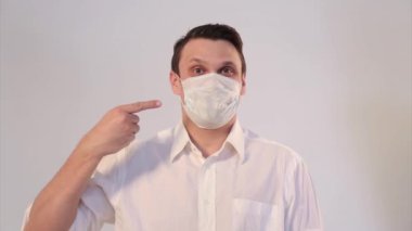 Beyaz arka planda tıbbi maske takan bir adamın portresi. Tıbbi ekipmanların tanıtımı. Adam işaret ediyor. Karantina, kendini izole etme, koronavirüsten koruma, virüs, hastalık korkusu, tedavi, ilaç