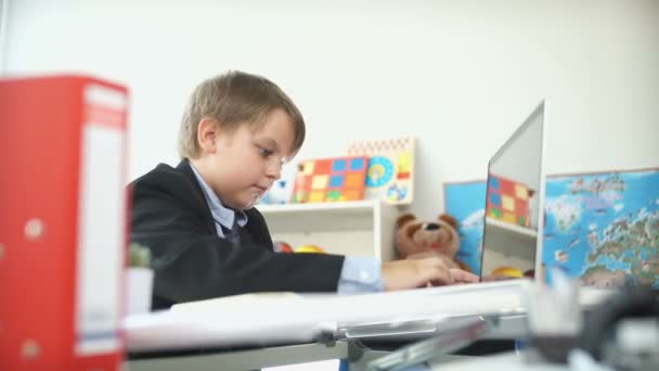 Αστείο μικρό αγόρι στο επιχειρηματικό κοστούμι και κάθεται στο τραπέζι δακτυλογραφούν στο φορητό υπολογιστή. — Αρχείο Βίντεο