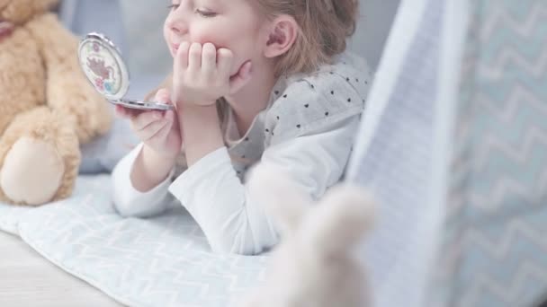 Маленькая девочка лежит на полу в детской, глядя в карманное зеркало — стоковое видео