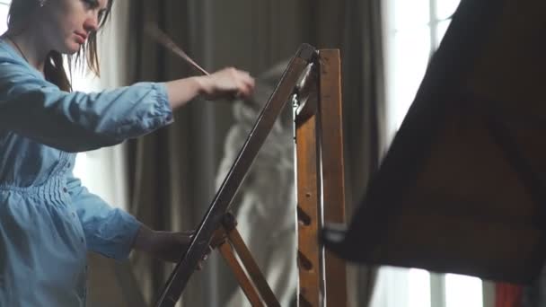 Young woman painter in blue dress paints on canvas in bright workshop — Stock Video