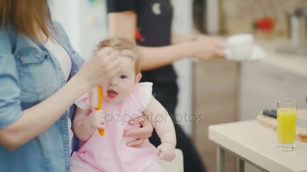 Маленькая милая девочка сидит на коленях у матерей и тянет щетку в рот — стоковое видео