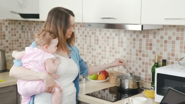 Mãe segura pequeno bebê nos braços e ao mesmo tempo prepara comida — Vídeo de Stock