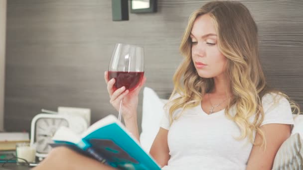 Bella giovane donna bionda che legge un libro e beve vino a letto — Video Stock