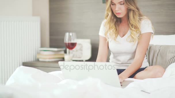 Frau in Hauskleidung sitzt auf einem Bett mit Laptop, Wein auf einem Tisch. — Stockvideo