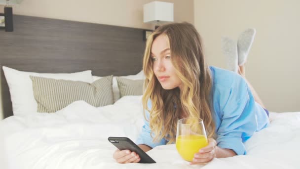 Γυναίκα βρίσκεται σε ένα κρεβάτι, ποτά χυμό πορτοκάλι, επικοινωνεί στο κινητό τηλέφωνο — Αρχείο Βίντεο