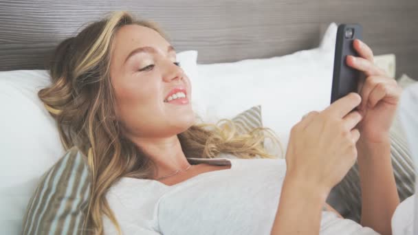 Beautiful woman in a light bed typing a message using a smartphone. — Stock Video