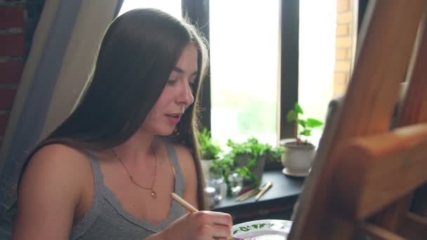 Длинноволосая девушка рисует с маслом цвета на мольберте в интерьере мастерской — стоковое видео