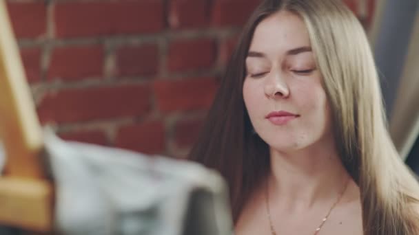 Long-haired girl paints with oil colors on easel in workshop interior. — Stock Video