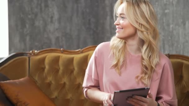 Happy young girl sitting on the sofa with tablet computer on gray back — Stock Video