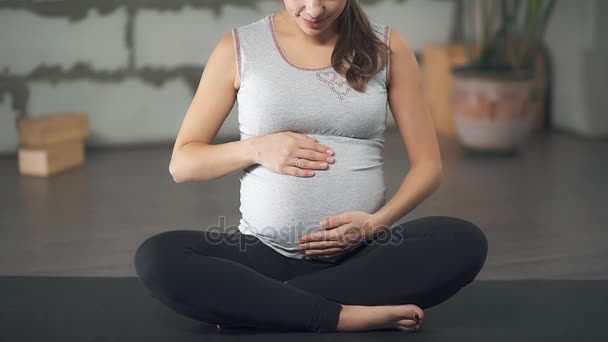 Una mujer embarazada está acariciando su vientre, sentada en una esterilla de yoga . — Vídeos de Stock