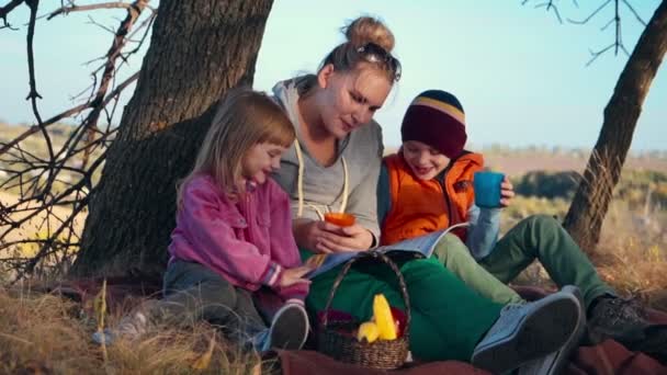 Mama i dzieci, ciesząc się ciepłym pić pod drzewem — Wideo stockowe