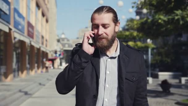 Skäggig man går längs en gata och prata via telefon — Stockvideo